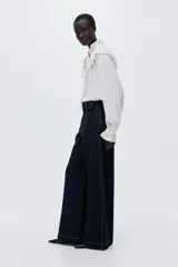 Pantalón de jean de tiro alto con pierna amplia y corte holgado. Confeccionado en denim rígido de color azul oscuro, presenta costuras decorativas verticales en el frente, bolsillos laterales y un cinturón integrado del mismo material con hebilla.