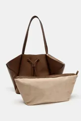 Bolso de hombro tipo maxi shopper color marrón, con doble asa y estructura flexible que se pliega hacia afuera. Presenta un detalle de tira ajustable con cordón en el centro frontal. Incluye un compartimento interior extraíble con cierre de cremallera y cierre principal mediante botón imantado.