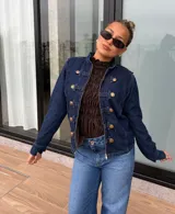 Chaqueta de denim azul oscuro con estilo militar, cuello alto, cierre frontal con cremallera y detalles de trabas decorativas con botones dorados.