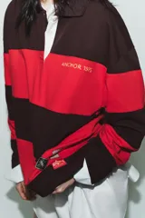 Polo de manga larga con cuello solapa y cierre frontal de botones. Diseño a rayas horizontales anchas en color negro y rojo. Presenta texto bordado en el pecho y parches decorativos en la parte inferior delantera.