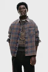 Camisa de franela de algodón suave con efecto lavado, corte relajado. Presenta cuello inglés, tapeta clásica de botones, un bolsillo superior abierto en el pecho y canesú en la espalda. Las mangas son largas con tapeta y puños abotonados, y el bajo es ligeramente redondeado. El estampado es a cuadros en tonos grises y naranjas.