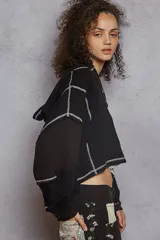 Buzo cropped de manga larga, color negro, con cuello tipo polo y cierre parcial de botones. Presenta un diseño de patchwork con paneles de tejido acanalado negro y un panel central de tela estampada con microflores blancas, todo unido con costuras expuestas en hilo blanco.