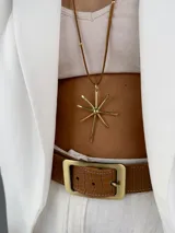 Colgante de diseño minimalista compuesto por una estructura circular rígida de metal dorado y un dije central en forma de estrella o destello de líneas rectas. Incluye tiras de cuero marrón con detalles metálicos que aportan un toque artesanal.