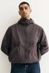 Campera rompevientos de nylon repelente al agua, color gris oscuro/marrón topo, con forro de malla. Presenta cierre frontal con protección para el mentón, capucha ajustable con cordón, bolsillos laterales y bolsillo interior con cierre, y elástico revestido en puños y bajo.