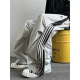 Pantalón deportivo unisex color beige, de corte ancho, con bolsillos laterales tipo cargo, cintura elástica ajustable con cordón, cierre lateral completo y detalle de tres rayas verticales negras en el lateral.