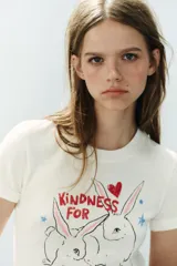Camiseta blanca de algodón con cuello redondo, manga corta y calce regular. Presenta un estampado frontal ilustrado con conejos, corazones, estrellas y el texto "Kindness for every kind".