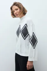 Media polera de tejido de punto color crudo, con diseño de rombos en el frente en tonos gris y negro. Presenta cuello alto y mangas largas.