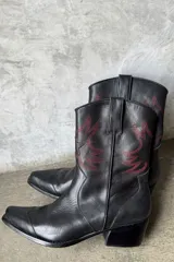 Botas tejanas de cuero negro con punta cuadrada y tacón bajo. Presentan bordados rojos de estilo western en la caña.
