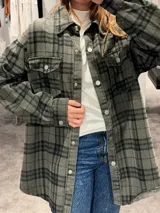 Camisa tipo sobrecamisa de corte oversize, confeccionada en tela a cuadros en tonos marrones y negros. Presenta cierre frontal con botones metálicos a presión, bolsillos frontales con solapa y mangas largas con puños abotonados.