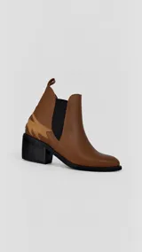 Botineta de cuero color marrón, estilo Chelsea, con inserciones elásticas laterales negras y detalle de aplicación de gamuza en el talón. Cuenta con taco cuadrado de madera y presilla trasera.