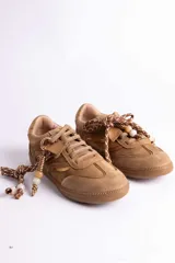 Zapatillas urbanas de gamuza color tostado, con diseño de cordones decorados con cuentas y detalles metálicos. Presentan suela de goma al tono y una estética inspirada en el estilo retro deportivo.