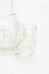 Jarra de vidrio transparente con diseño de estrellas doradas. Incluye un vaso de vidrio transparente con diseño de líneas verticales doradas.