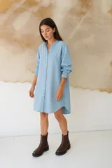 Vestido camisero de denim azul claro, con cuello mao, manga larga con elástico en el puño y cierre frontal de botones. El ruedo es asimétrico con corte en V en la parte delantera.