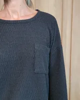 Remerón de textura acanalada en color gris claro, con cuello redondo, mangas largas y bolsillo frontal en el pecho. Presenta un diseño holgado con corte asimétrico, siendo más largo en la parte posterior.