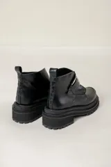 Bota corta de cuero negro con suela track dentada y taco cuadrado. Presenta una tira decorativa en el empeine con una hebilla metálica plateada y tirador en la parte trasera.