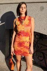 Vestido corto sin mangas con cuello redondo, confeccionado en microtul con un estampado en tonos cálidos como rojo, naranja y amarillo que simula un efecto tie-dye. Presenta un diseño ajustado al cuerpo con detalle de drapeado lateral en la cintura.