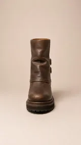 Bota de caña media confeccionada en cuero marrón con acabado desgastado. Presenta un diseño de solapa lateral con dos correas ajustables con hebillas metálicas y suela de goma dentada con plataforma.