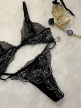 Conjunto de lencería compuesto por bralette triangular y tanga, confeccionado en encaje bitono negro y blanco. El bralette presenta cierre frontal con broche de gancho y breteles regulables. La tanga es de estilo colaless con laterales regulables mediante herrajes metálicos.