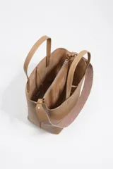 Bolso de hombro tipo tote bag de piel vacuna color beige, con doble asa de mano y correa ajustable de tejido. Presenta un cierre de cordón en la parte superior y un logo metálico en relieve en el frente. Incluye bolsillo interior con cierre de cremallera.