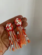 Caravanas artesanales con base superior de resina en color marrón con detalles de lámina dorada y cristales incrustados. Presentan flecos largos de hilo marrón combinados con tiras de strass brillante.