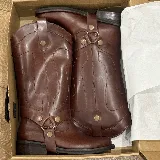 Bota de caña alta estilo western en cuero color marrón, con detalle de arnés con tachas metálicas y tiradores laterales.