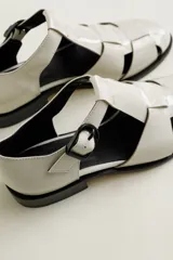 Sandalia cerrada tipo pescador, color blanco o hueso brillante, con tiras entrecruzadas en el empeine y cierre de hebilla ajustable en el lateral. Posee suela y taco bajo de color negro.