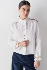 Camisa blanca de lino con cuello alto tipo gorguera, cierre frontal con botones de madera y detalles de ribetes en color mostaza en hombros y puños. Presenta volados en la botonera, cuello y puños.