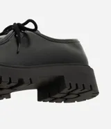 Zapato estilo desert shoe u oxford de Satinato, confeccionado en poliuretano (PU) negro con acabado texturizado. Presenta diseño de punta redonda, cordones al tono y suela gruesa tractorada de goma.