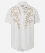 Camisa de corte comfort confeccionada en algodón, color blanco, con cuello solapa, manga corta y cierre frontal de botones. Presenta un delicado bordado floral en tono beige en la parte superior delantera.