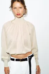 Blusa marrón de punto fluido, con cuello subido abotonado en un lateral y mangas largas con puños ajustados. Presenta un diseño fruncido en la parte frontal que aporta volumen.
