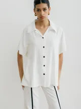 Camisa blanca de manga corta, corte amplio y cuello tipo solapa. Cierre frontal con botones negros.