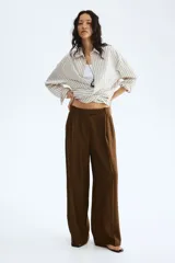 Pantalón sastrero de corte ancho, color marrón, con pinzas frontales, trabillas para cinturón y raya marcada en las piernas.