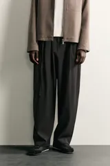 Pantalón de corte balloon fit en color gris oscuro, confeccionado en tejido con viscosa. Presenta pliegues frontales, cintura ajustable con cinturón integrado, bolsillos laterales y bolsillos traseros de doble vivo. Cierre frontal con cremallera y botón.