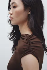 Top sin mangas color marrón oscuro, de punto elástico, con ajuste entallado. Presenta un adorno de metal y una tira larga en un hombro que forma un escote asimétrico. Los laterales están fruncidos para crear un efecto drapeado.