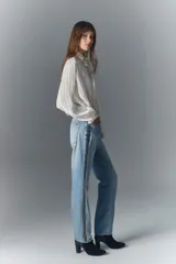Jean de tiro alto confeccionado en denim azul rígido con lavado claro localizado y efecto de bigotes. Presenta un corte de pierna recta y apliques laterales de tachas brillantes.