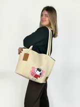 Tote bag de cuero color natural con diseño intervenido a mano por la artista GGIL, presentando a Snoopy con gafas de sol y corona. Incluye un sobre a juego con el mismo diseño artístico. Posee doble asa de hombro y diseño sin forro.