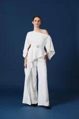 Mono largo de color blanco con diseño de un solo hombro y mangas amplias tipo capa. Presenta un corte peplum en la cintura con un cinturón integrado que incluye una hebilla metálica dorada, y pantalón de corte recto y fluido.