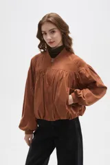 Chaqueta tipo bomber color tostado, confeccionada en material efecto gamuza. Presenta diseño con frunces en el pecho y espalda, cuello mao, cierre frontal completo y mangas largas con puños elásticos.