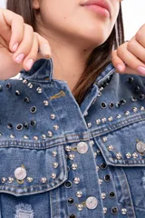 Campera de jean cropped con tachas plateadas distribuidas en el frente, hombros y mangas. Presenta un diseño con bordes deshilachados, cuello clásico y cierre frontal con botones metálicos.