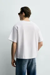 Camiseta de corte boxy fit, color blanco, con cuello redondo y manga corta. Presenta una aplicación de etiquetas decorativas a contraste en el centro del pecho.