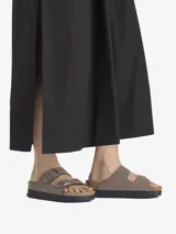 Sandalia tipo slide de dos tiras ajustables, color marrón topo, con hebillas metálicas color oro rosa. Presenta una plataforma gruesa de color negro sobre la clásica suela de corcho anatómica de Birkenstock.