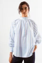 Blusa de algodón con diseño a rayas verticales azules sobre fondo blanco. Presenta cuello alto con volados, cierre frontal con botones pequeños y mangas largas abullonadas con puños ajustados.