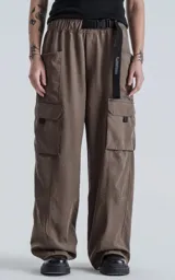 Pantalón cargo de silueta oversize, color marrón oscuro, confeccionado en tejido técnico liviano de poliéster. Presenta cintura elástica y múltiples bolsillos funcionales tipo cargo en las piernas. Incluye un cinturón de cinta negra con hebilla de plástico.