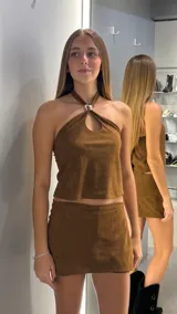 Top de gamuza color marrón chocolate, con escote halter y detalle de argolla metálica en el centro del pecho. Presenta un diseño con abertura tipo gota y corte recto.