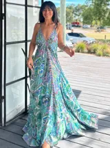 Vestido largo tipo solera, con escote en V profundo y tirantes finos. Presenta un estampado paisley en tonos rosados, lilas y blancos, con ajuste de lazo en la cintura y volados en el ruedo.