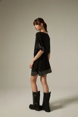 Vestido corto de poliéster color negro con escote a la base, mangas cortas y diseño holgado. Cuenta con detalles de tachas metálicas plateadas distribuidas por la prenda y agrupadas en el dobladillo y los puños.