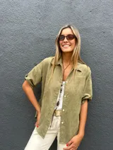 Camisa de gamuza sintética liviana en color verde oliva, con cuello clásico, cierre frontal de botones y mangas cortas con dobladillo.