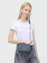 Bandolera Marc Jacobs modelo The Quilted Denim Softshot, confeccionada en denim azul acolchado con patrón de rombos. Presenta el logo Double J en metal plateado, correa de cuero y cadena, y cierre de cremallera doble.