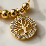 Collar de acero con baño de oro, compuesto por una cadena fina con esferas decorativas y un dije circular con el diseño del árbol de la vida rodeado de cristales.