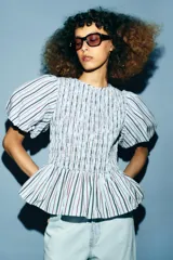 Top de cuello redondo con cuerpo de nido de abeja elástico y mangas cortas abullonadas con elástico en los puños. Presenta un diseño de rayas verticales en tonos azul, rojo y negro, con un bajo estilo peplum y cierre de botón en la espalda.